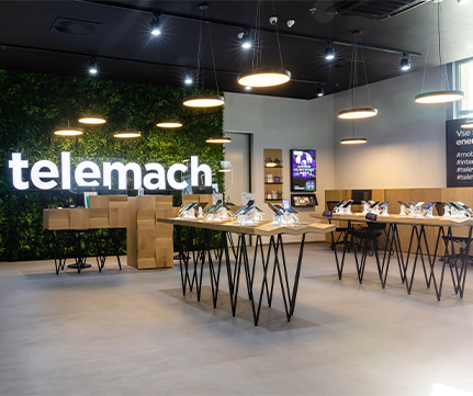 Moderna trgovina Telemach s svetlimi tlemi, razstavljenimi telefoni in recepcijo z velikim osvetljenim logotipom. Oblika trgovine je svetla in sodobna.