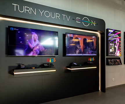 Sodoben televizijski salon ima dva elegantna zaslona, ki prikazujeta živahne vizualne elemente pod napisom TURN YOUR TV EON in poudarjata napredno zabavno tehnologijo.