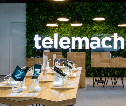 Sodobna notranjost trgovine Telemach z elegantnimi lesenimi mizami, na katerih so razstavljeni pametni telefoni in tablični računalniki, osvetljeni s toplimi visečimi lučmi ob bujni zeleni steni.